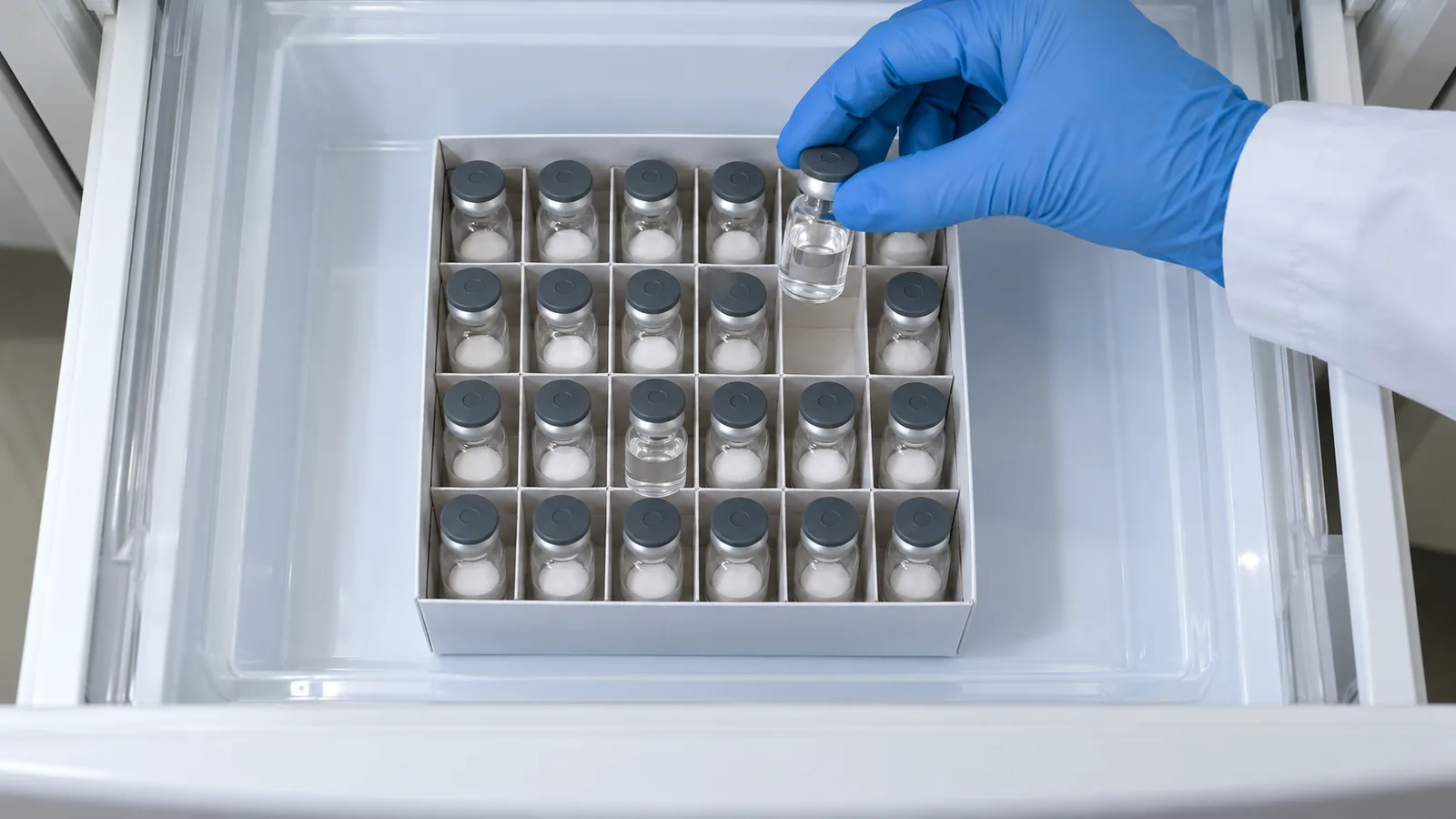Gloved hand lifting a vial of clear mixed tirzepatide from a box of powder vials in a laboratory refrigerator drawer